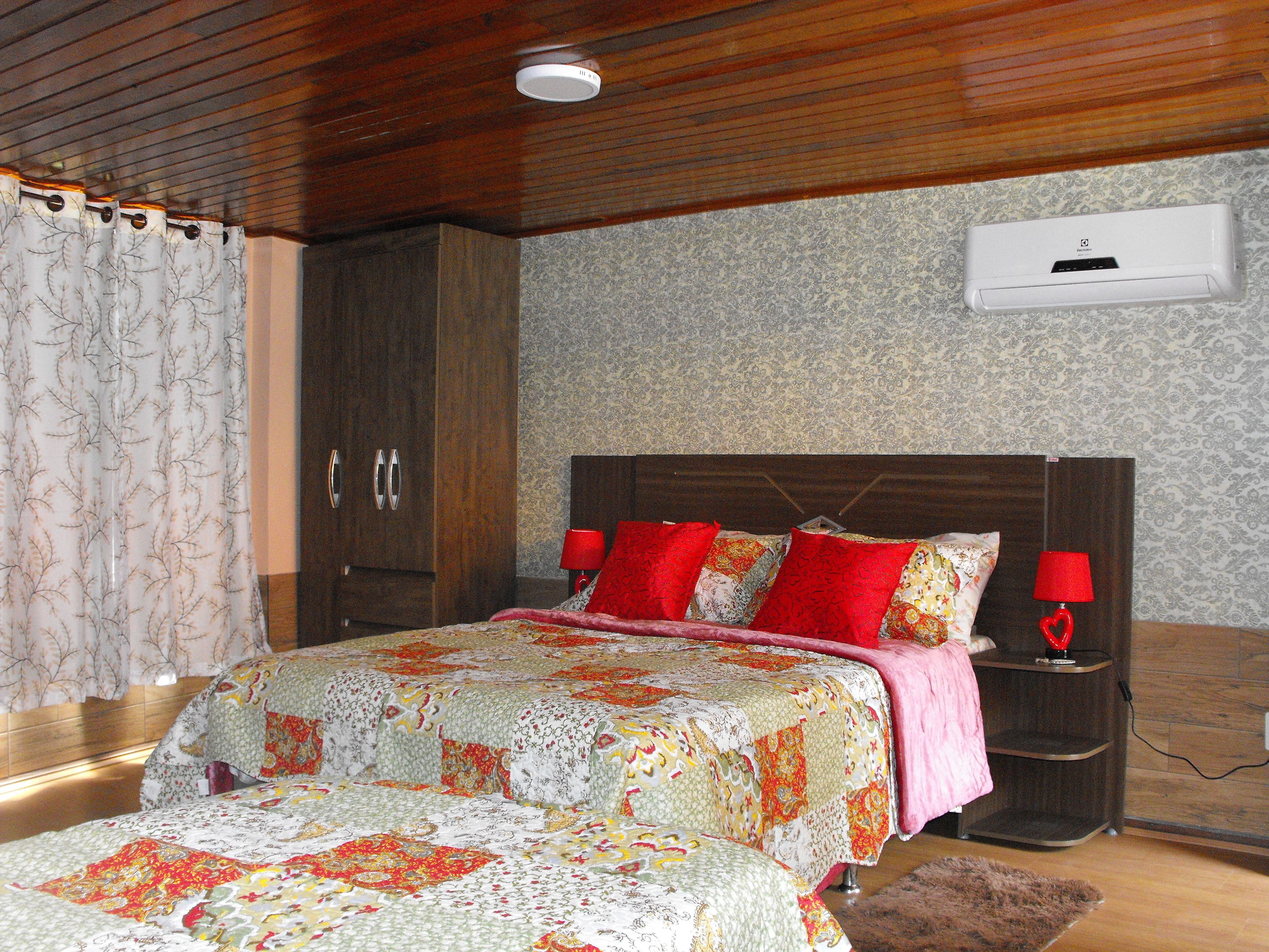 Un dormitorio con techo de madera, una cama con colcha, dos mesitas de noche con lámparas y un aire acondicionado de pared.