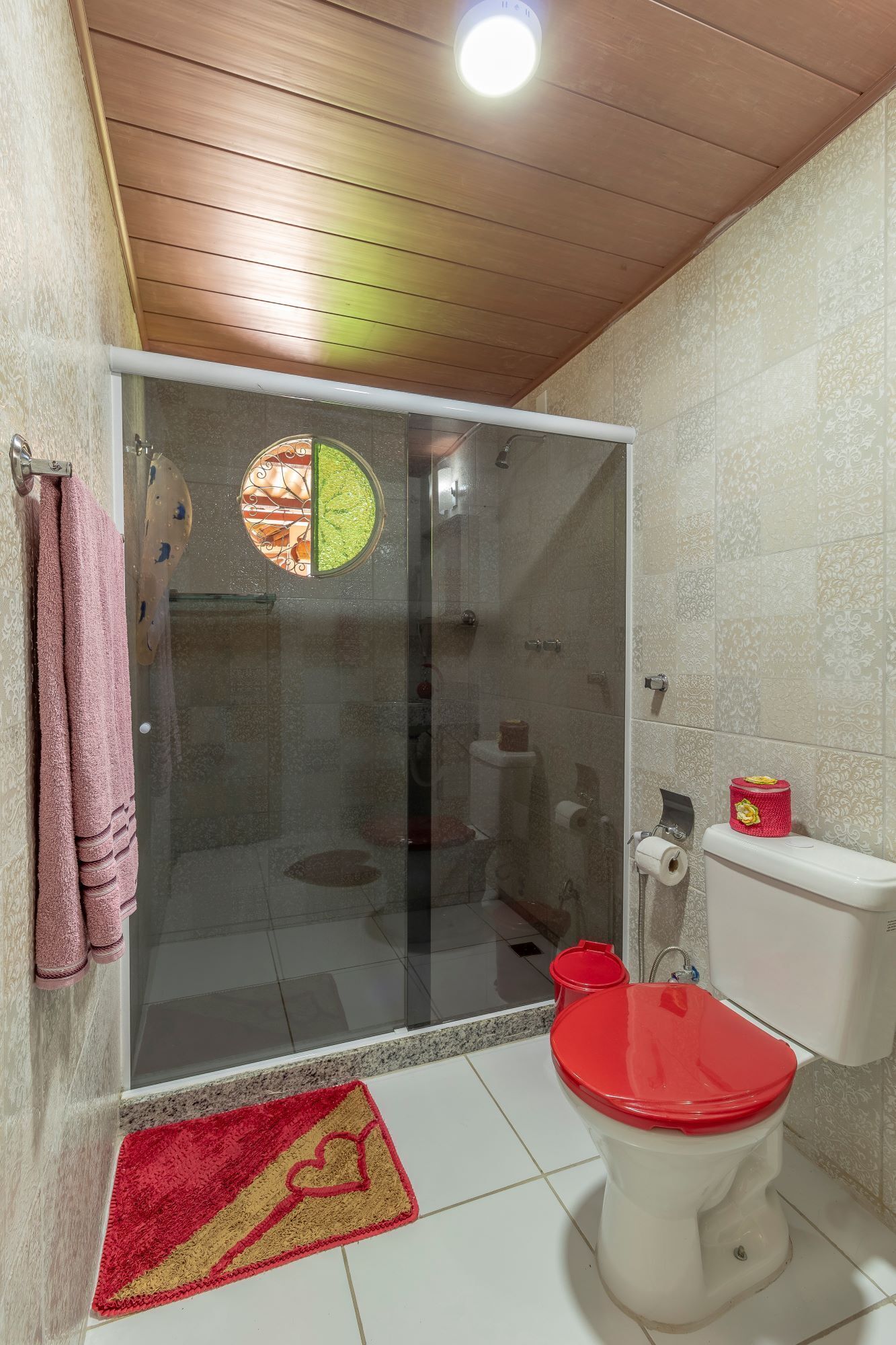 Un baño con cabina de ducha de vidrio, inodoro con asiento rojo y alfombra estampada en el suelo.
