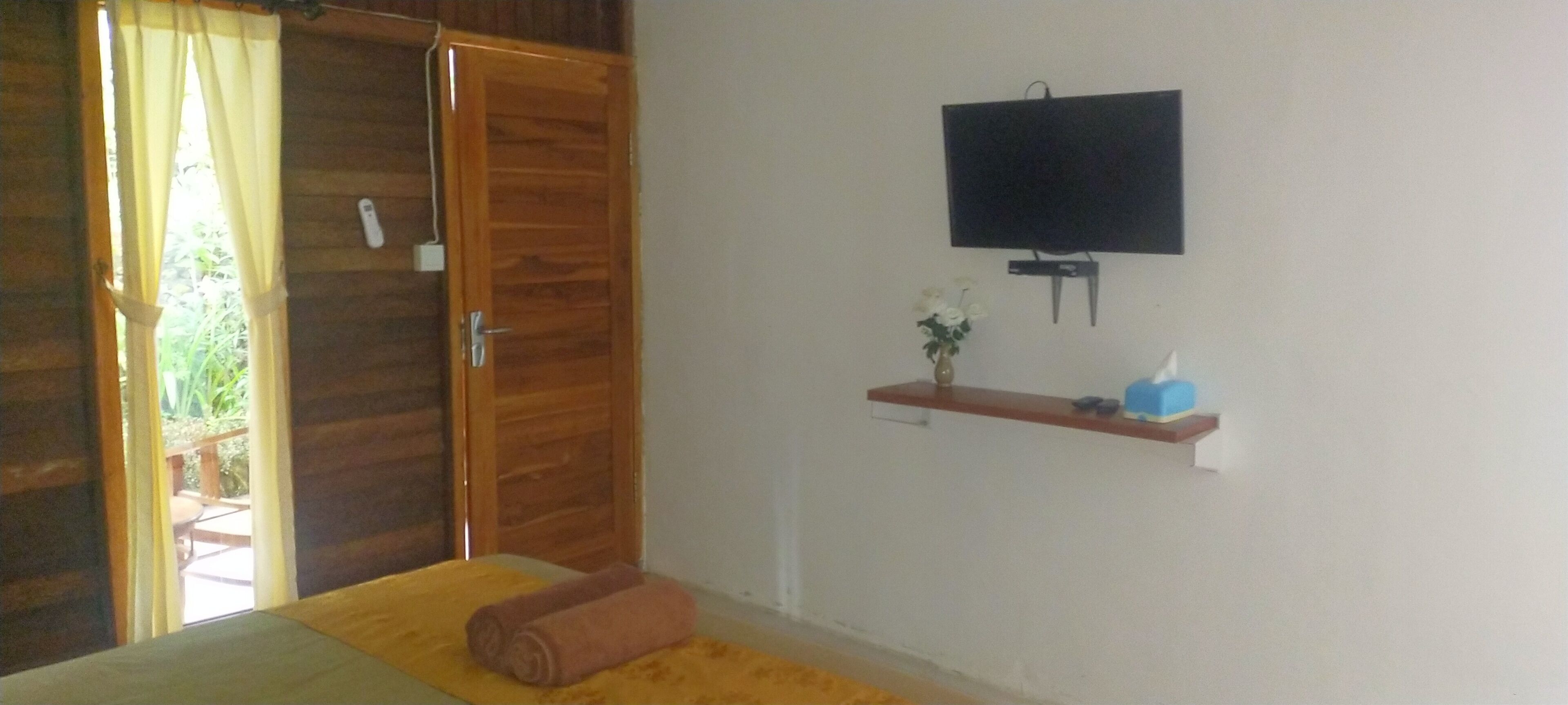A room with a wooden door, a bed with a yellow bedspread, a TV mounted on the wall, and a small shelf with a vase and a blue object.