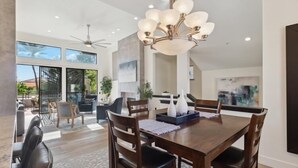 A spacious dining area with a wooden table and chairs, a modern chandelier, and large windows offering views of the outdoors. - Comfort in a Condo Gainey Ranch Style (Scottsdale)