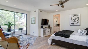 A modern bedroom with a large bed, a TV, a ceiling fan, and a view of palm trees through the window. - Comfort in a Condo Gainey Ranch Style (Scottsdale)