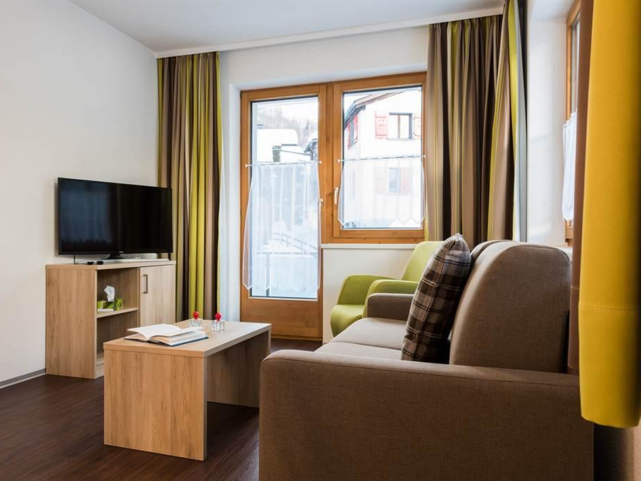 Ein modernes Wohnzimmer mit Flachbildfernseher, Couchtisch aus Holz, einer Couch und einem Fenster mit Vorhängen.
