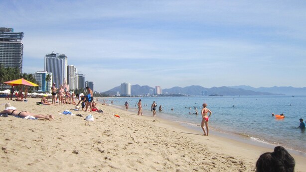 A beach with people sunbathing and swimming, a cityscape with tall buildings in the background, and a clear sky. - Forget Me Not Hotel Nha Trang (Nha Trang)