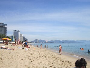 Een strand met mensen die zonnebaden en zwemmen, een stadsbeeld met hoge gebouwen op de achtergrond, en een heldere lucht.