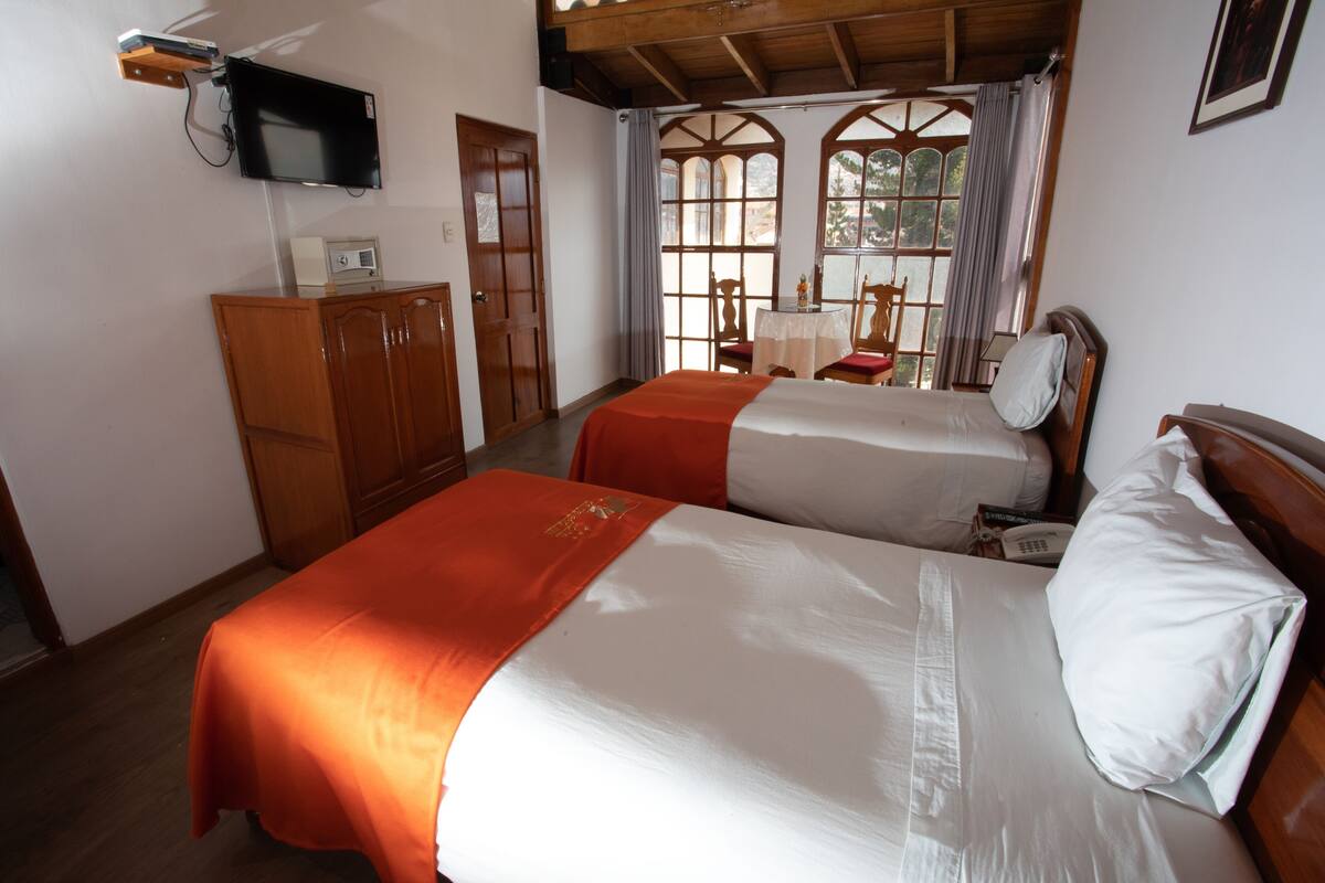 Ein Hotelzimmer mit zwei Betten, einem an der Wand befestigten Fernseher, einem Schreibtisch mit Stuhl und einem Vorhangfenster.