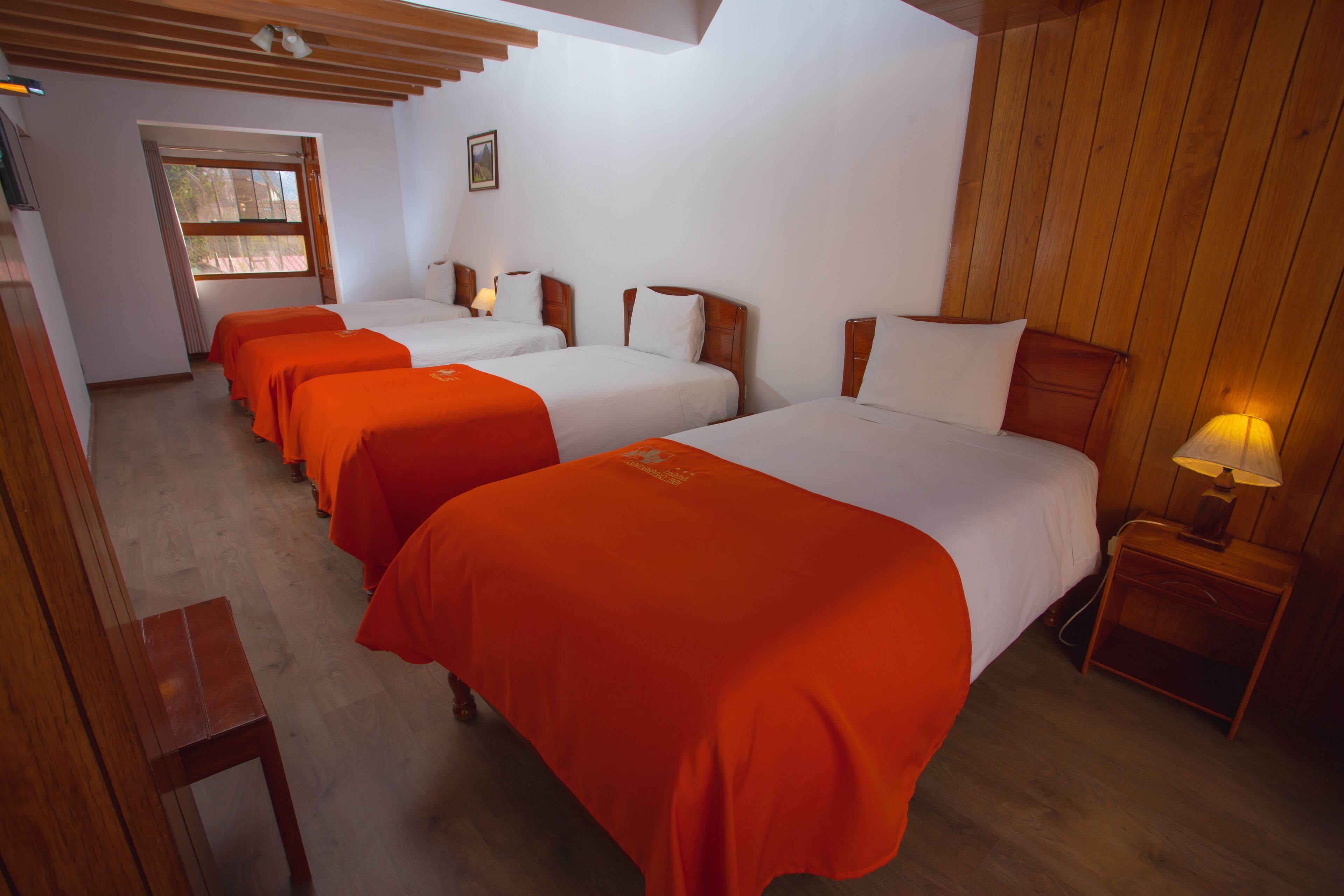 Una camera d'albergo con quattro letti singoli, poggiateste in legno e un comodino con una lampada.