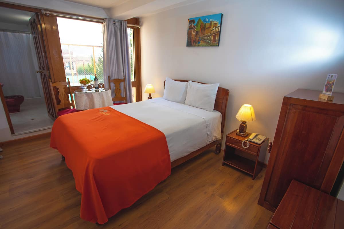 Ein Hotelzimmer mit Bett, Nachttischen aus Holz, einem Tisch mit Lampe, einem Bild an der Wand und Blick durch ein Fenster.