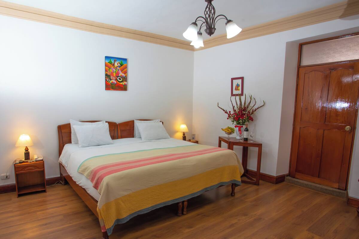Ein ordentlich bezogenes Bett mit farbenfroher Bettdecke, ein Holz-Kopfbrett, Nachttischlampen, ein Bild an der Wand, ein kleiner Tisch mit Blumenvase und eine Holztür.