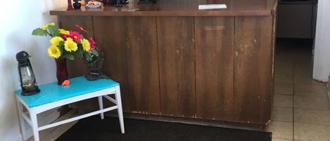 A reception area with a wooden desk, a small white bench, a vase of flowers, a lamp, and a cross on the wall.