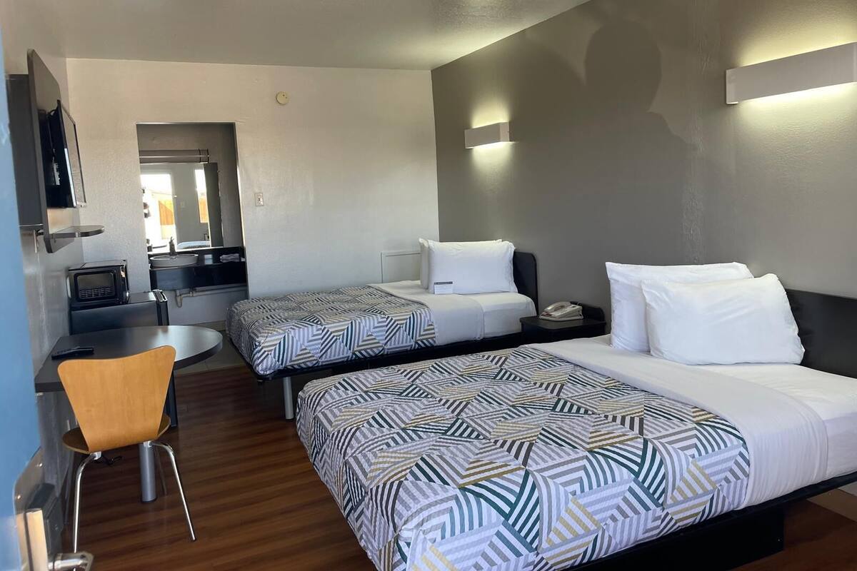 A hotel room with two beds, a small desk, a chair, a TV, and a kitchenette.