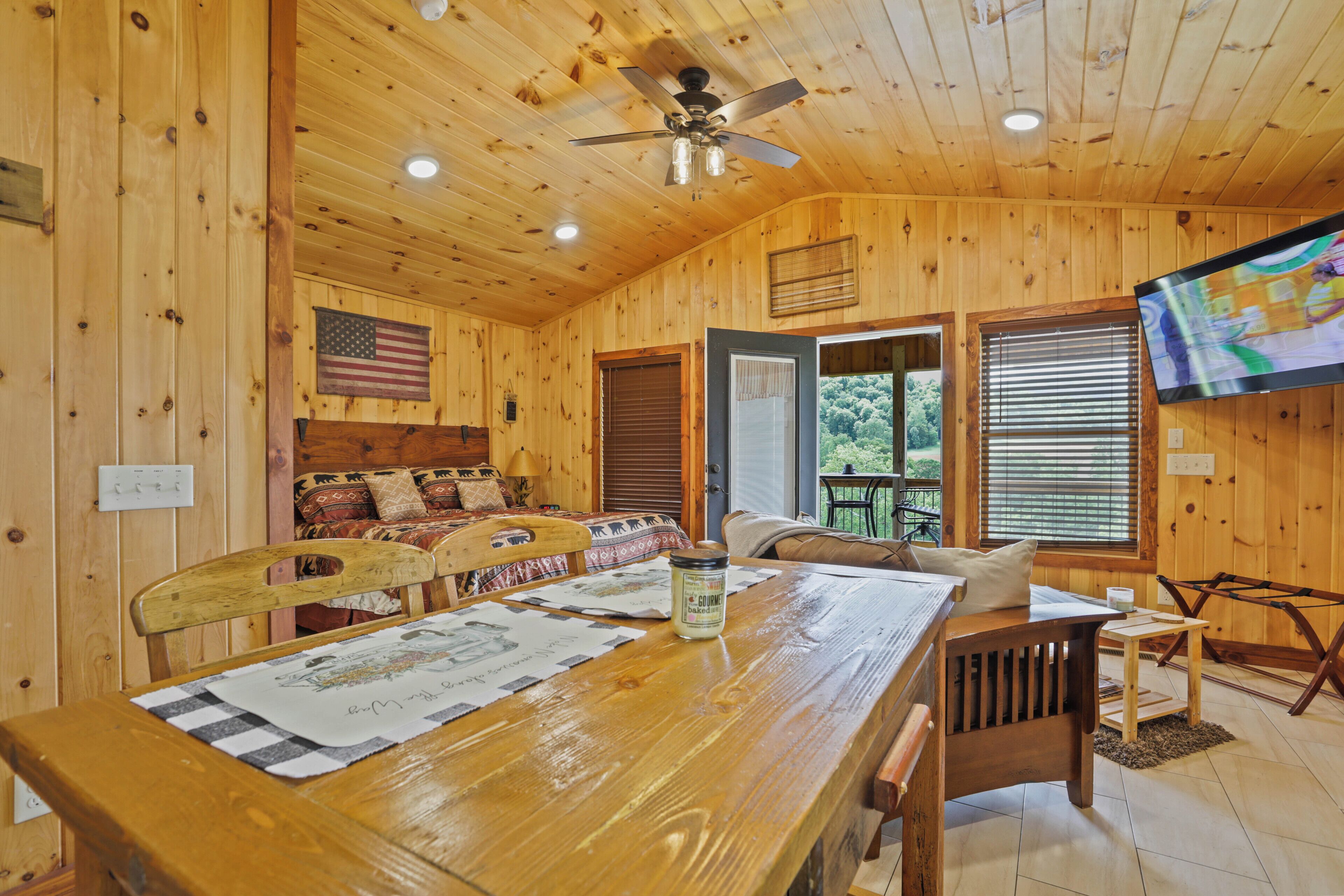 A wooden cabin interior with a dining table, chairs, a ceiling fan, and a television mounted on the wall.