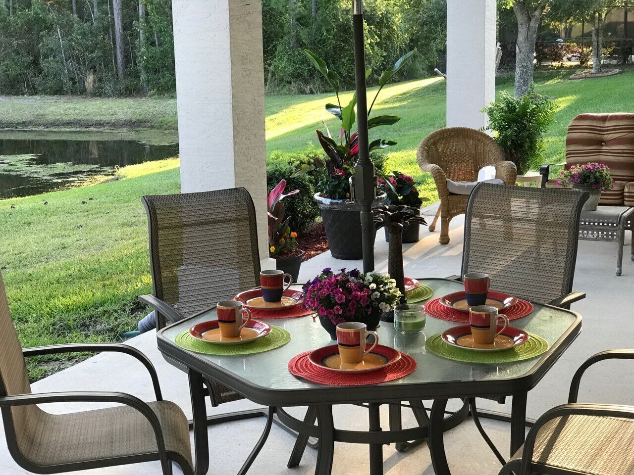 An outdoor patio with a table set for four, surrounded by wicker chairs, with a view of a pond and a deer in the background.