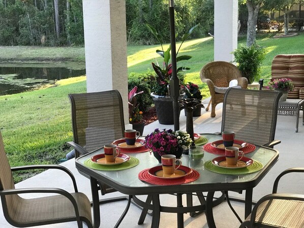 An outdoor patio with a table set for four, surrounded by wicker chairs, with a view of a pond and a deer in the background.