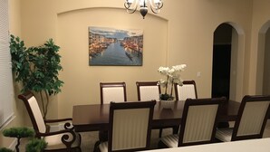A dining room with a table set for six, surrounded by chairs, a chandelier, a painting of a canal, and potted plants.