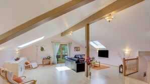 Un loft spacieux avec de hauts plafonds, des poutres apparentes, un espace de vie comprenant un canapé et une table basse, un coin repas et un escalier offrant une vue sur l’extérieur.