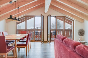 Une pièce spacieuse avec un plafond haut, des poutres en bois, une table à manger avec des chaises, un canapé rouge et de grandes portes vitrées donnant sur un espace extérieur.