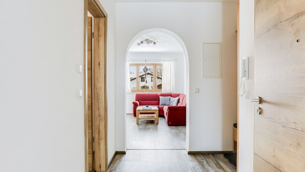 Een hal die uitkomt op een woonkamer met een rode bank, houten vloeren en een uitzicht door een rondboogvormige doorgang.