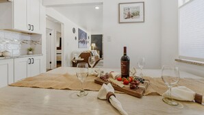 Moderni keittiö, jossa on valkoiset kaapit, ruokailutila puupöydän kera, ja taustalla olohuone.