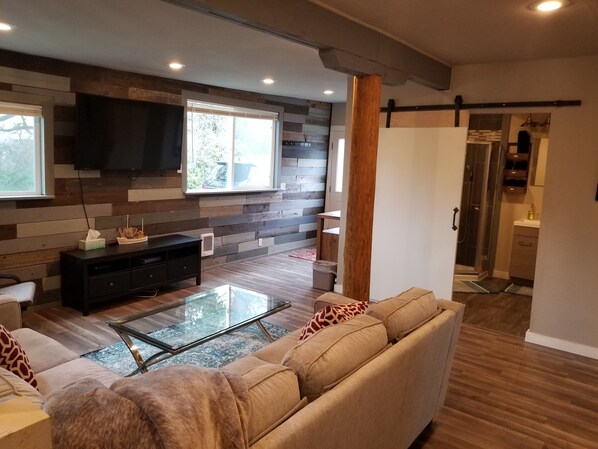 A modern living room with a sofa, a glass coffee table, and a TV mounted on a wooden wall.