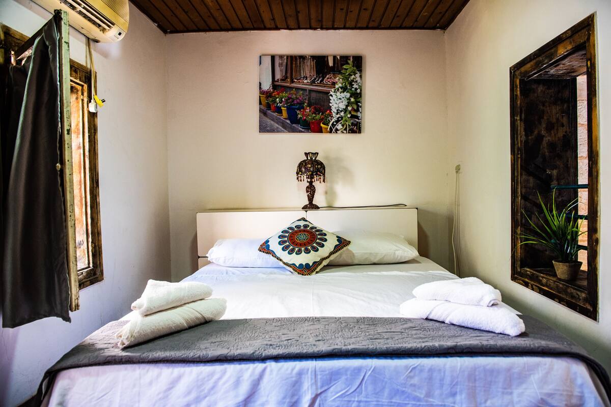 A neatly made bed with white linens and a decorative pillow, a wall-mounted air conditioner, a framed picture of flowers, and a window with a curtain.