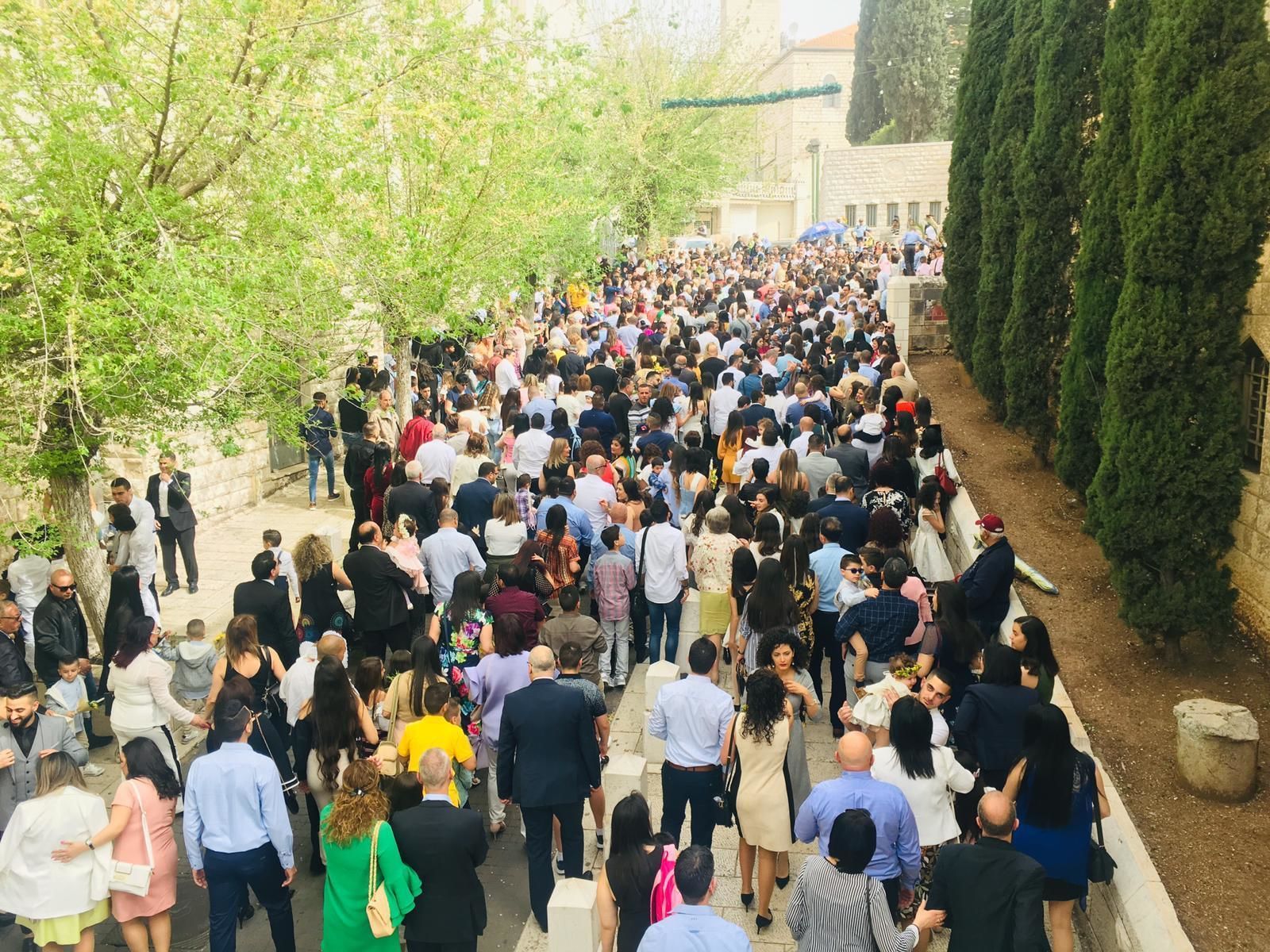 A crowded outdoor area with people walking and standing, surrounded by trees and a stone building.