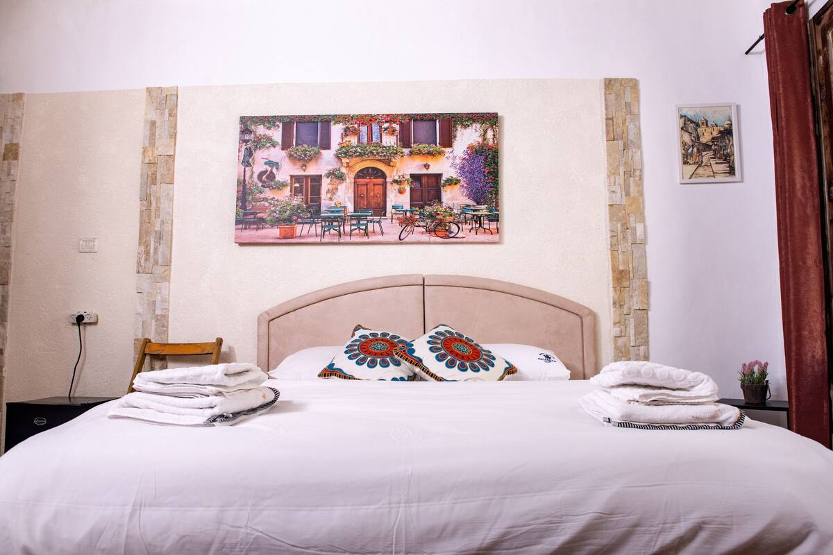 A hotel room with a bed, a headboard, a painting of a street scene, and a small table with a plant.