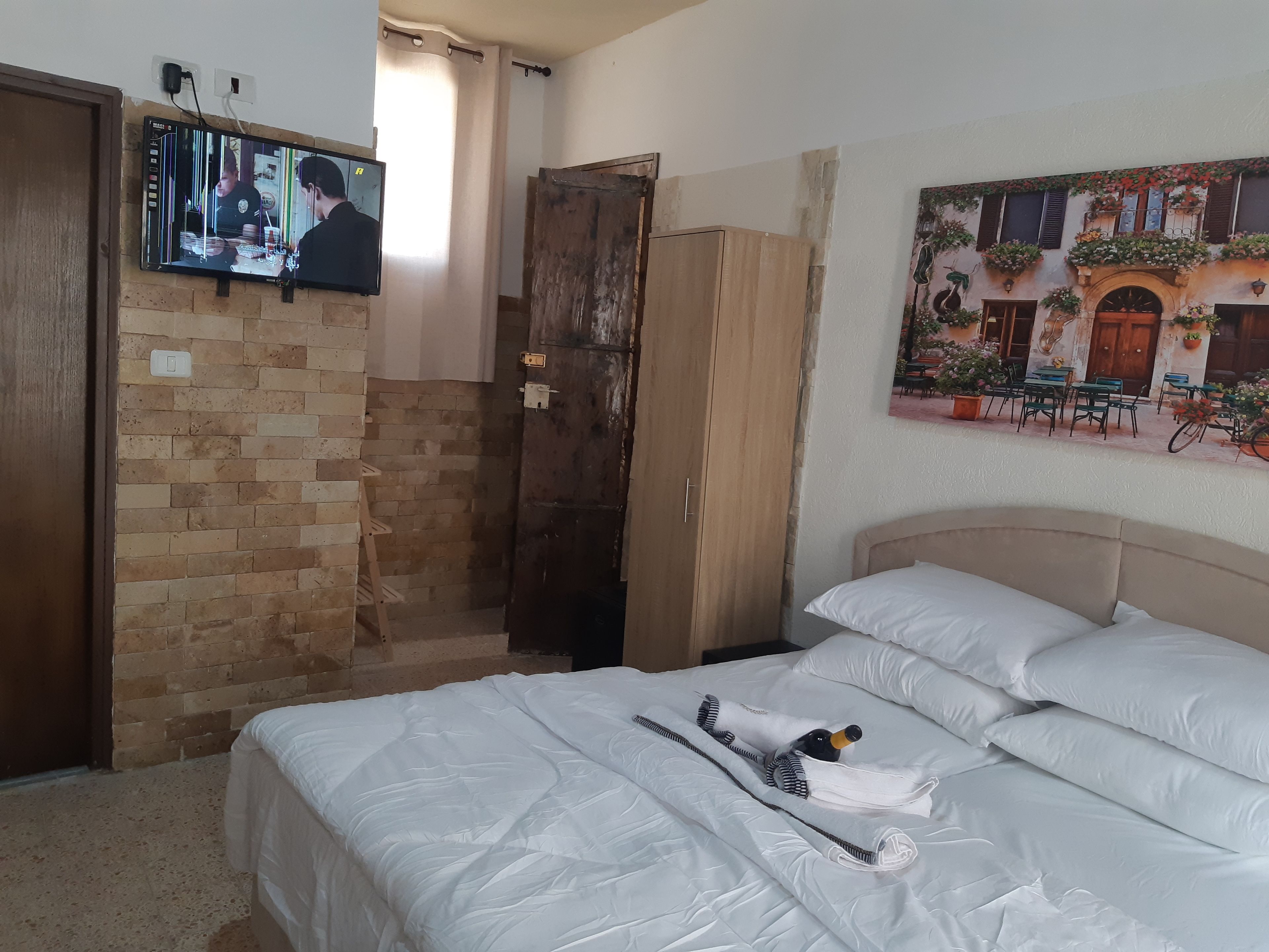 A hotel room with a brick wall, a wooden wardrobe, a bed with white bedding, and a TV mounted on the wall.