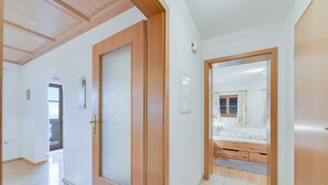 Un pasillo con puertas de madera, suelo de baldosas y una habitación visible con cama y ventana.