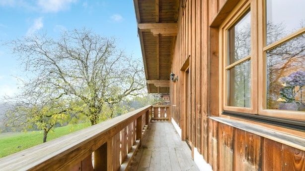Un balcón de madera con barandilla, ventanas amplias y vistas a árboles y un campo cubierto de césped.