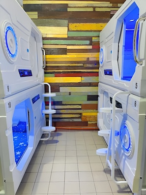 A row of white, modern-looking, multi-functional units with blue lighting, numbered 5 to 10, against a colorful, wooden panel wall. - Zzz Sleep Mode Capsule (Johor Bahru)