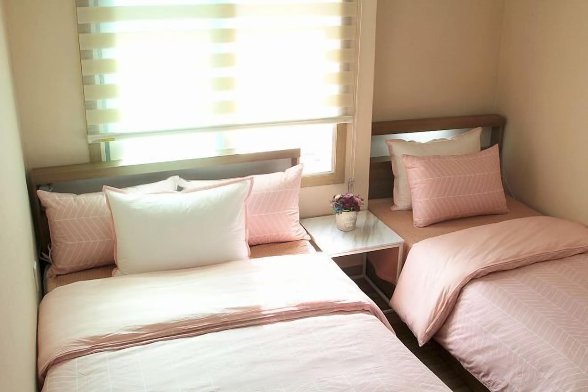 A hotel room with two beds, each with pink bedding and pillows, a small table with a vase of flowers, and a window with blinds.