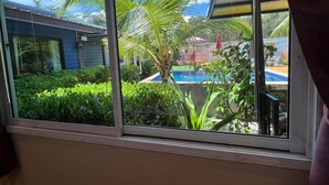 A window with a view of a swimming pool surrounded by lush greenery and a house in the background. - At Thara Aonang (Krabi)
