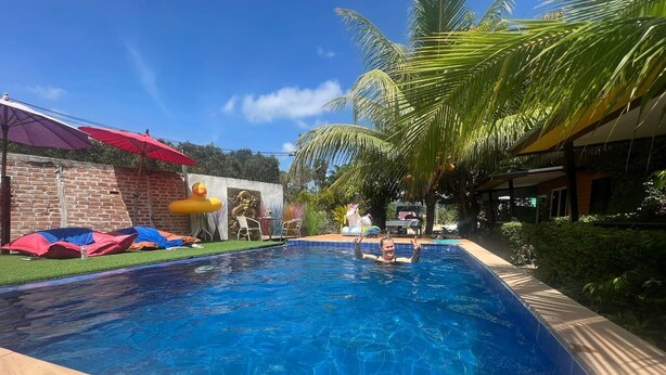 A swimming pool with clear blue water, surrounded by a tiled deck. There are colorful umbrellas and lounge chairs on the left side. - At Thara Aonang (Krabi)