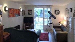 A living room with a sofa, a television, a telescope, and a dining area with a table and chairs. - Charming T2 garden with trees and parking, close to beaches and golf course. (Seignosse)