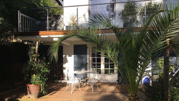 A patio with a white table and chairs, surrounded by potted plants and a wooden deck. - Charming T2 garden with trees and parking, close to beaches and golf course. (Seignosse)