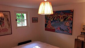 A bedroom with a bed, a window, a lamp, and a colorful painting on the wall. - Charming T2 garden with trees and parking, close to beaches and golf course. (Seignosse)