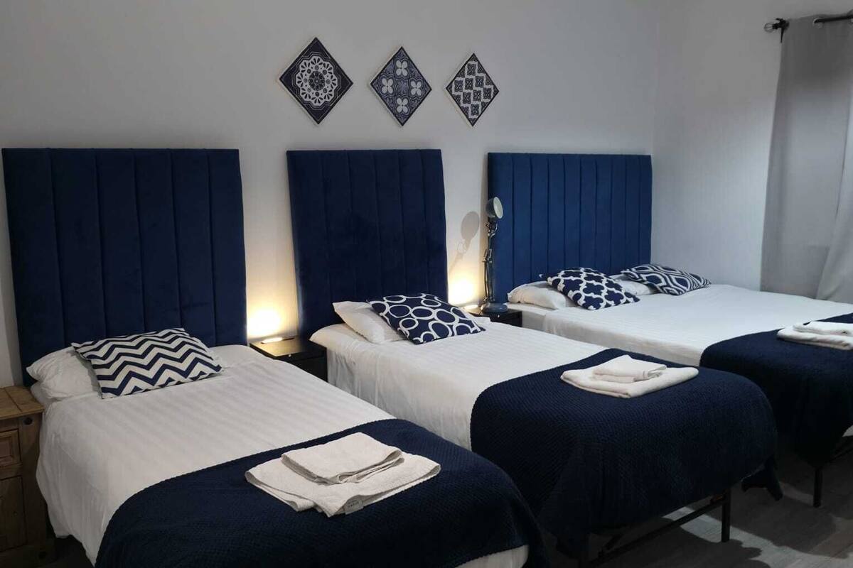 A hotel room with three beds, each with a blue headboard and white bedding. There are decorative wall tiles above the beds and a ceiling light fixture.