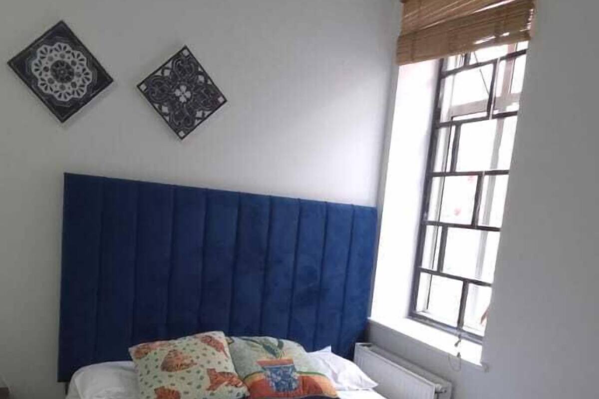 A bedroom with a large bed, a blue headboard, two decorative wall pieces, a window with blinds, and a radiator.