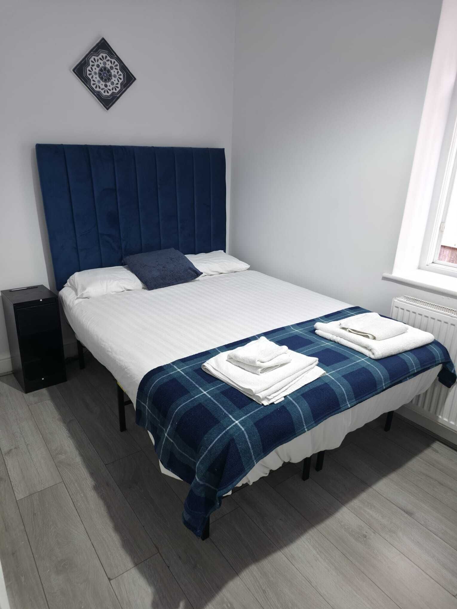 A neatly made bed with a blue headboard, white bedding, and a blue and white checkered blanket. There is a black speaker on the floor to the left of the bed.