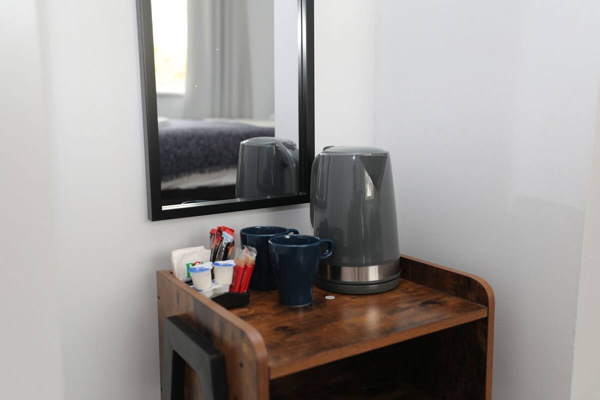 A small wooden table with a coffee maker, cups, and a mirror reflecting a bed and window.