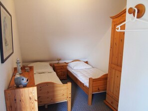 Une petite chambre à coucher accueillante, avec deux lits en bois, une commode en bois, un petit bureau avec une lampe et un tableau encadré au mur.