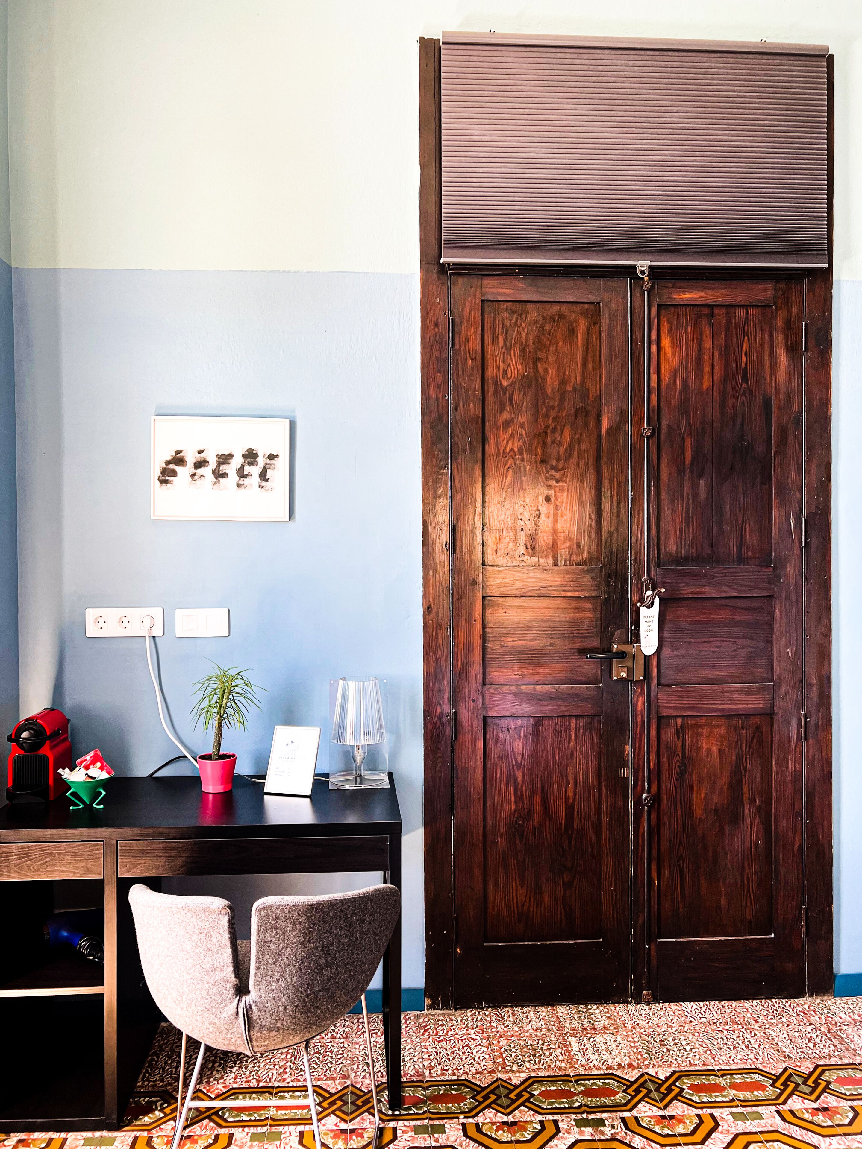 A room with a wooden desk, a chair, a potted plant, a lamp, and a large wooden door.