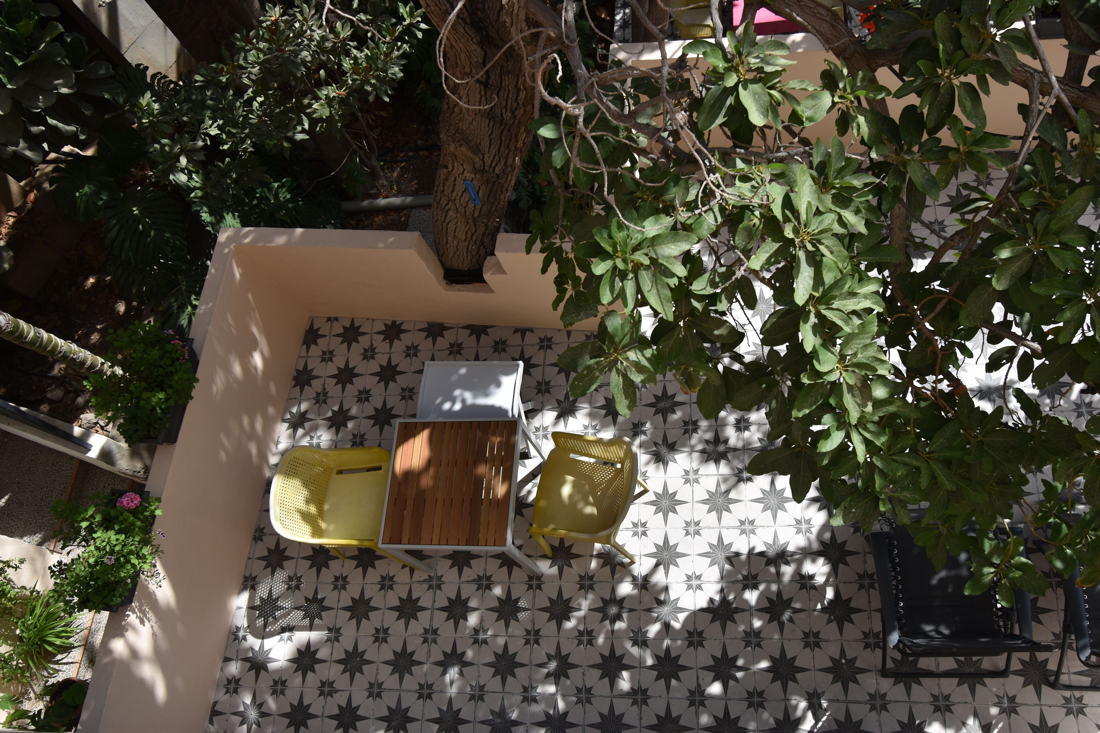 Un patio con suelo de baldosas, una mesa de madera, dos sillas y un árbol que da sombra.