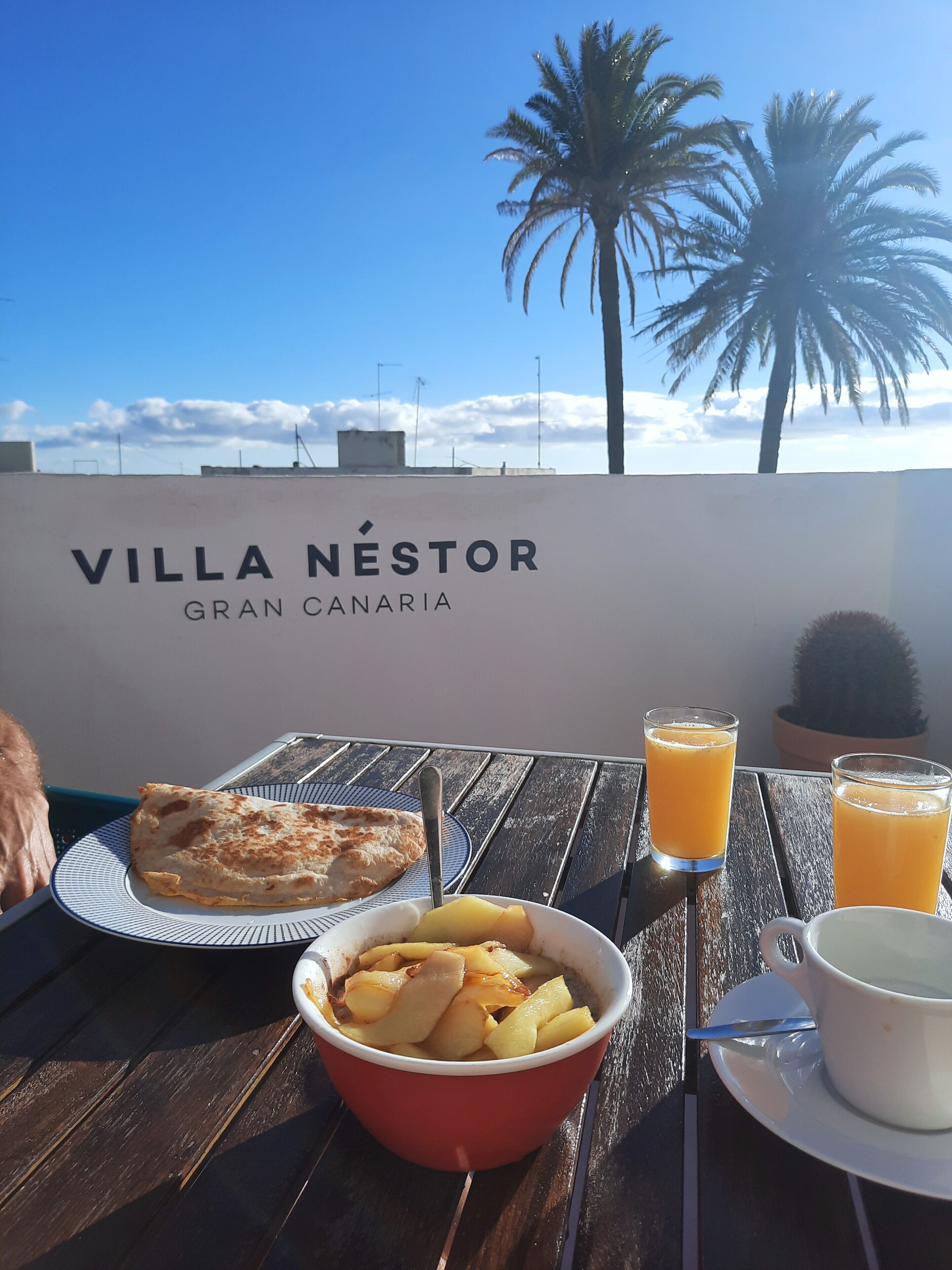 A table with breakfast items including a plate of food, a bowl of fruit, and two glasses of orange juice, with a view of the sky and palm trees in the background.