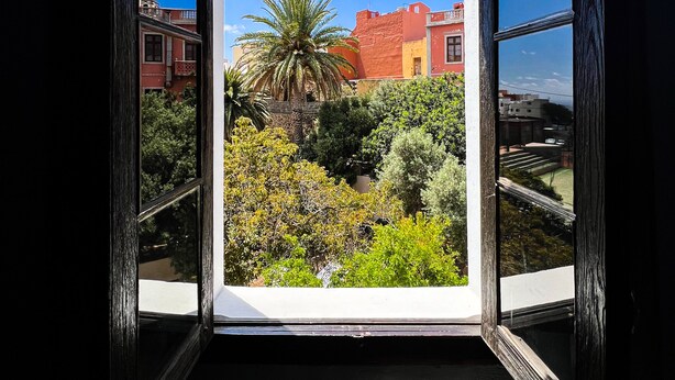 A room with a large window revealing a view of a garden with various plants and a building in the background. - Villa Nestor (Ingenio)
