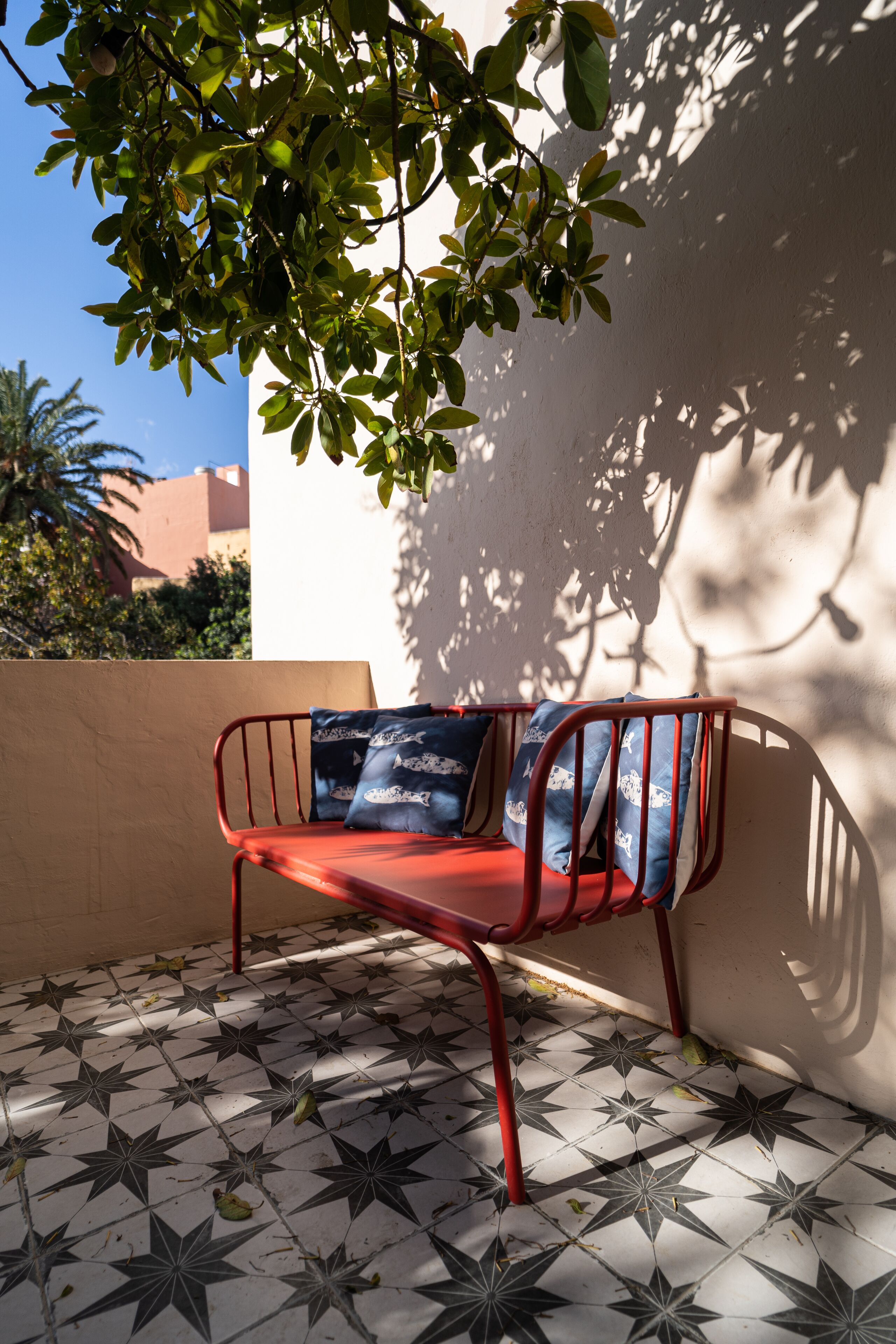 Un banc en métal rouge avec des coussins bleus, adossé à un mur blanc, sur un sol carrelé, et un arbre qui projette des ombres.