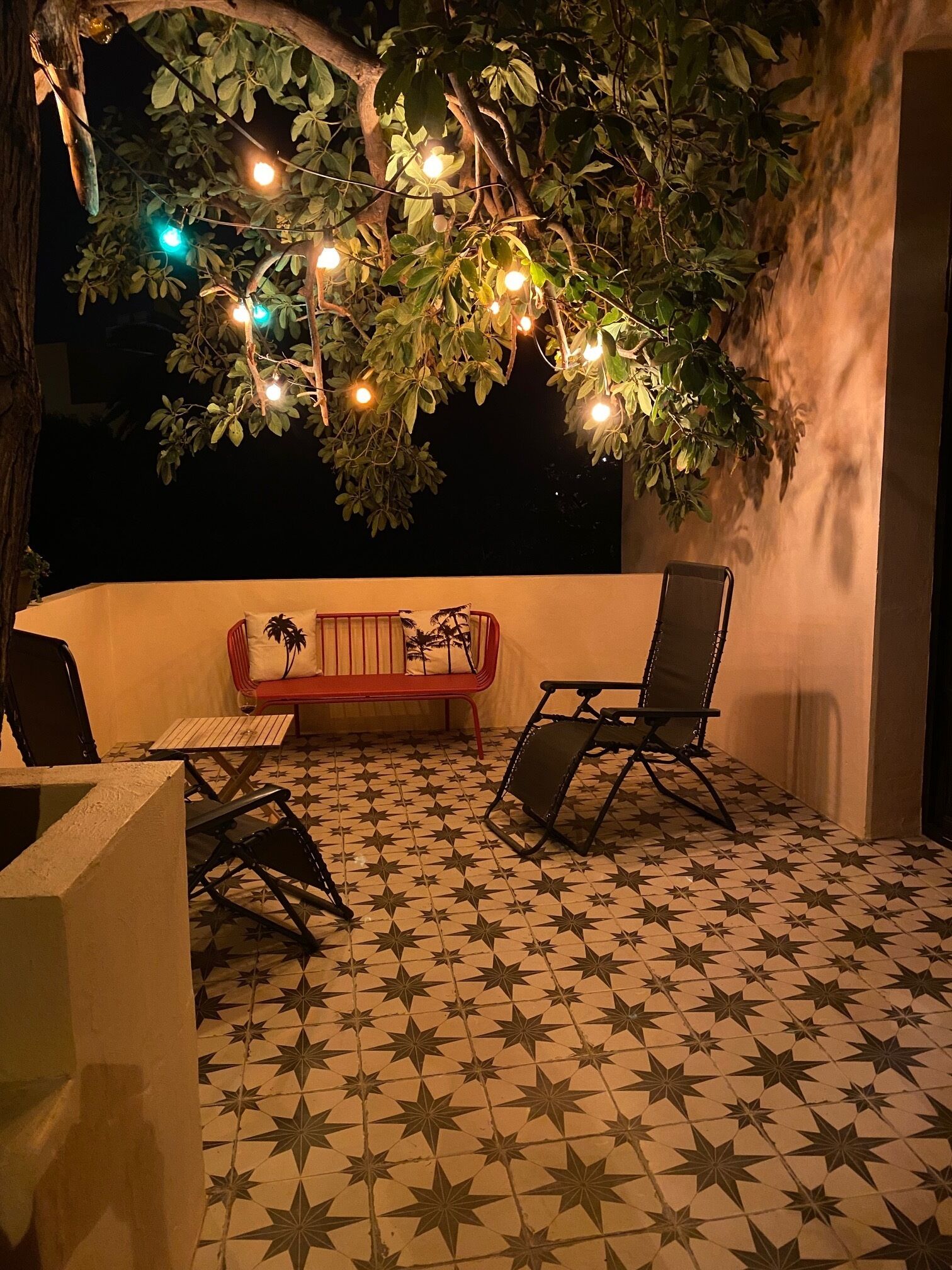 An outdoor patio with a star-patterned tiled floor, a red sofa, a black folding chair, and string lights hanging from a tree.