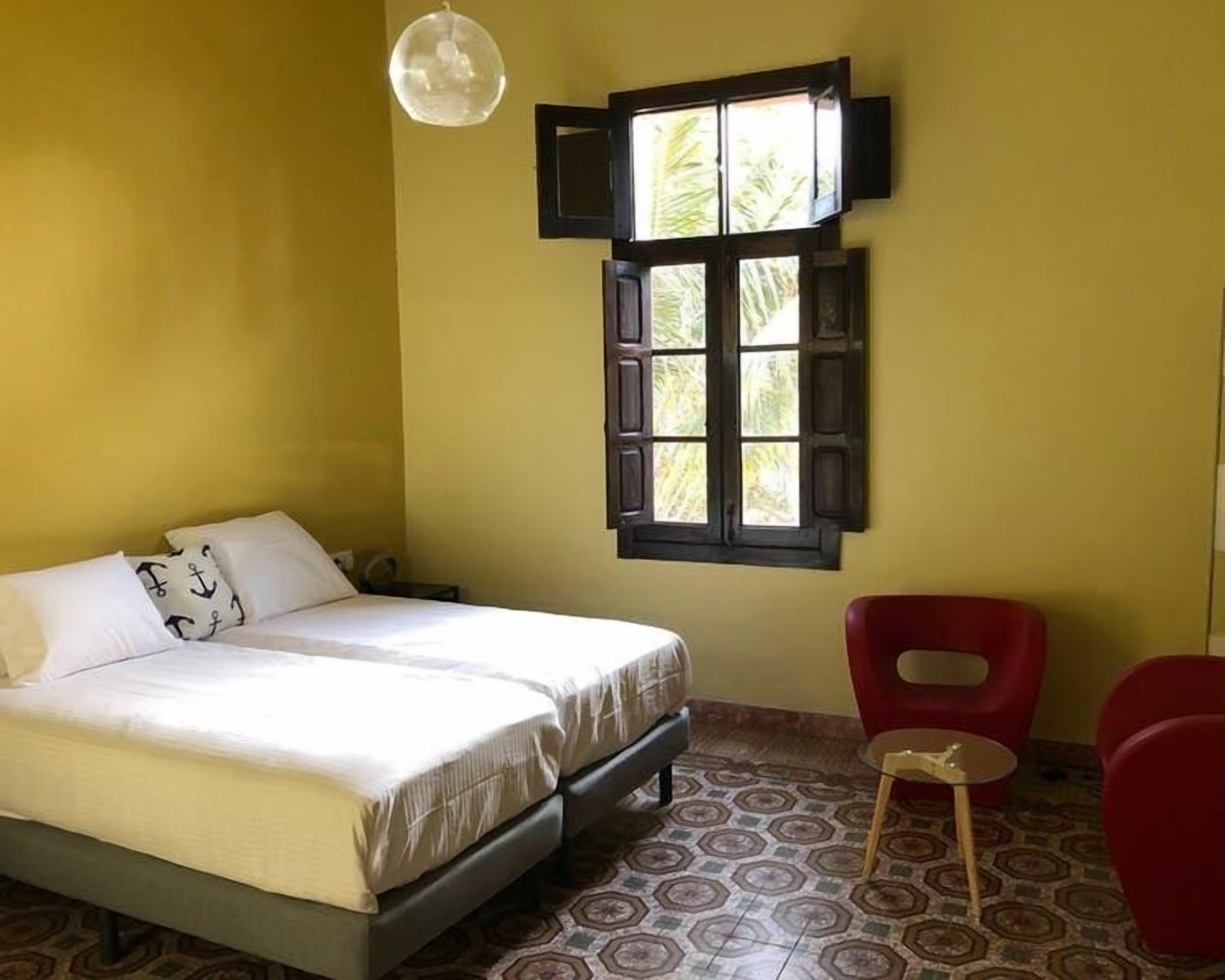 A room with a bed, a window with shutters, a red chair, and patterned flooring.
