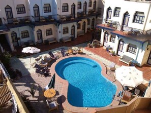 Una piscina con sillones de descanso y sombrillas, rodeada por un patio con edificios.