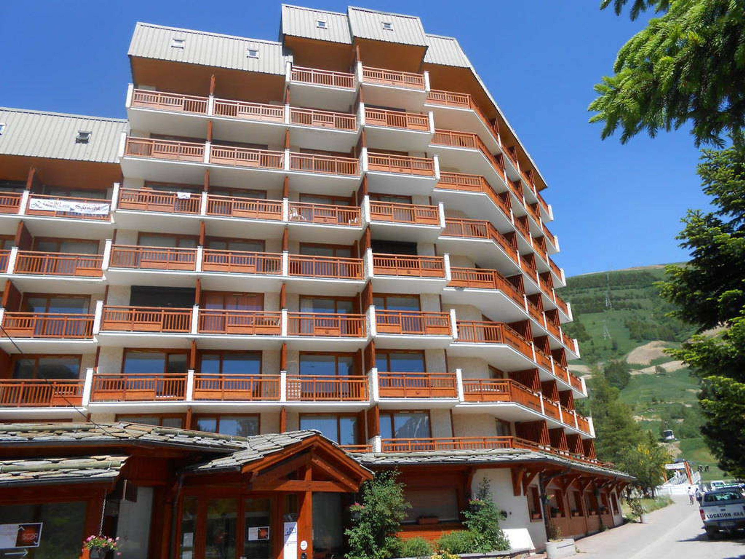 A multi-story hotel with balconies, a ski resort in the background, and a clear blue sky.